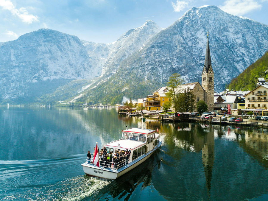 A passenger tour boat cruising on Lake Hallstatt toward the picturesque village of Hallstatt, Austria, with its church steeple and Alpine mountains in the background